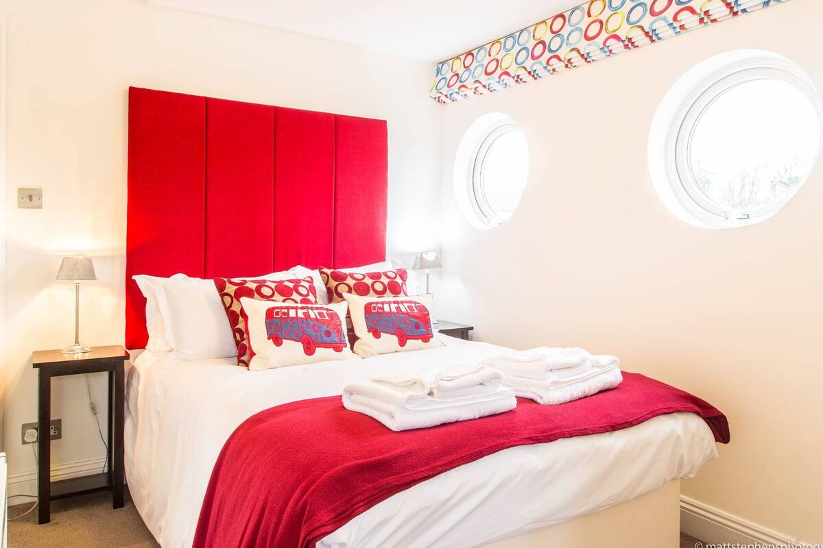 A modern bedroom with a red headboard, white bedding, and circular windows.