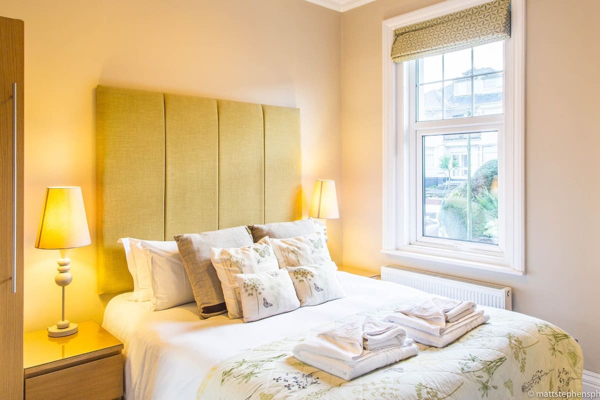 A neatly made bed with a green headboard, two bedside lamps, and a window with a view of buildings and greenery.