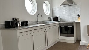 A modern kitchen with white cabinetry, a stainless steel range hood, a built-in oven, and two round windows providing natural light.