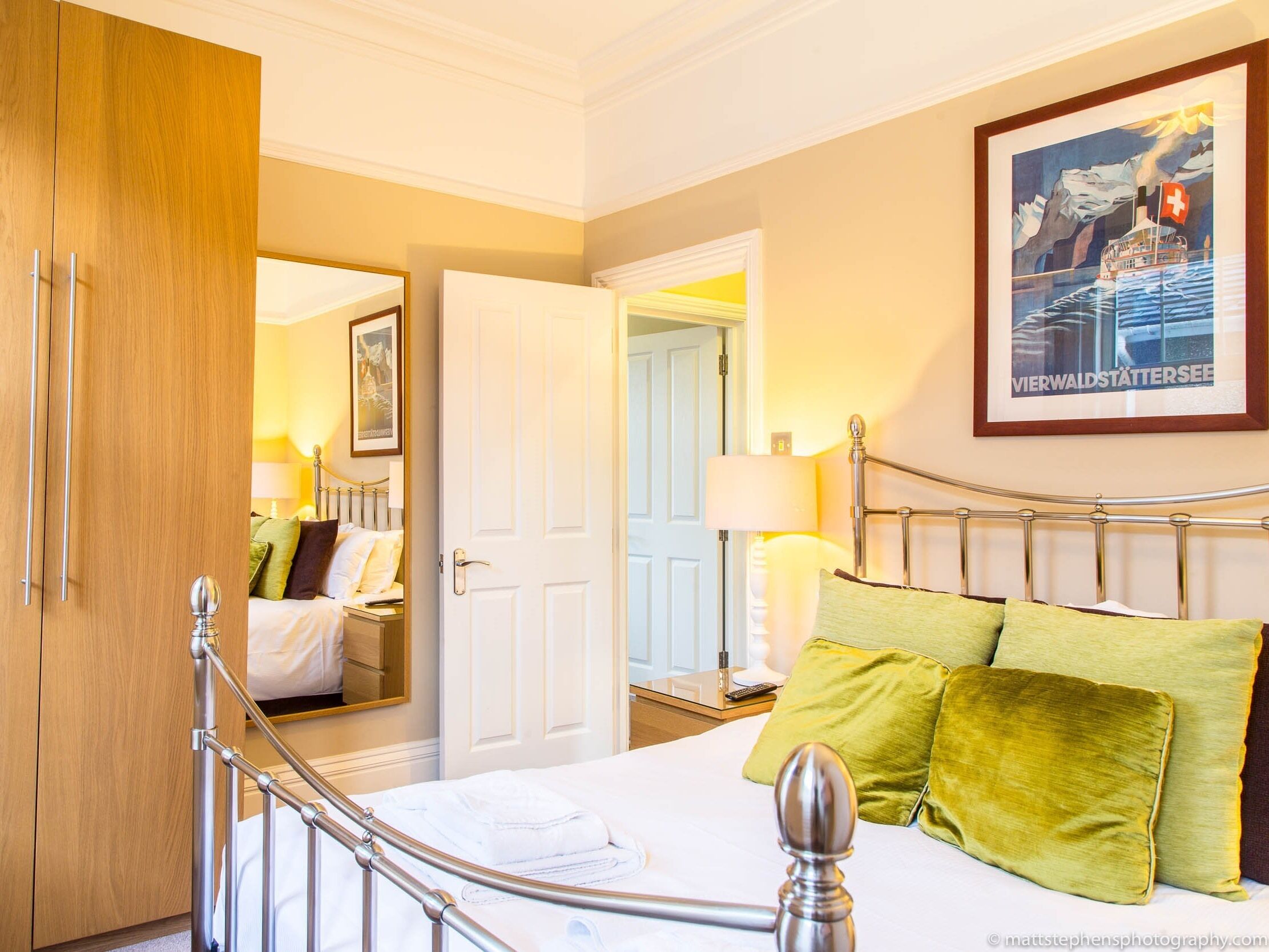 A hotel room with a bed, a wooden wardrobe, a bedside table with a lamp, and a framed picture on the wall.