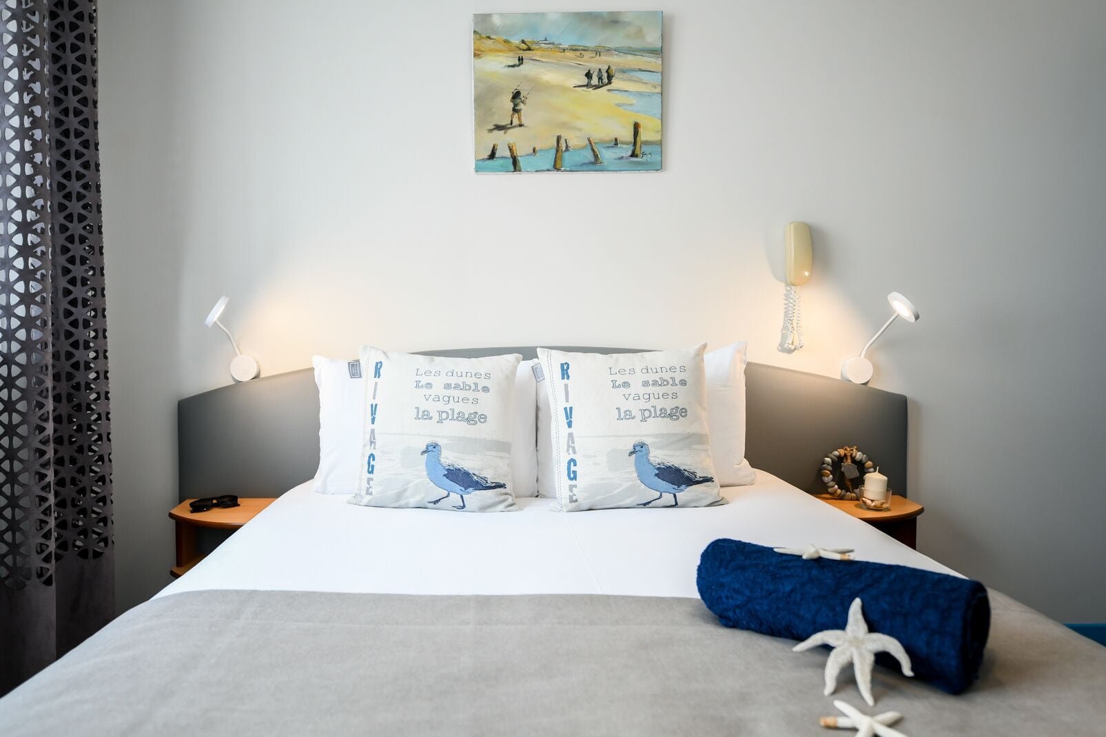 A neatly made bed with two pillows featuring a bird and text, a bedside lamp, a painting on the wall, and a starfish decoration on the bed.