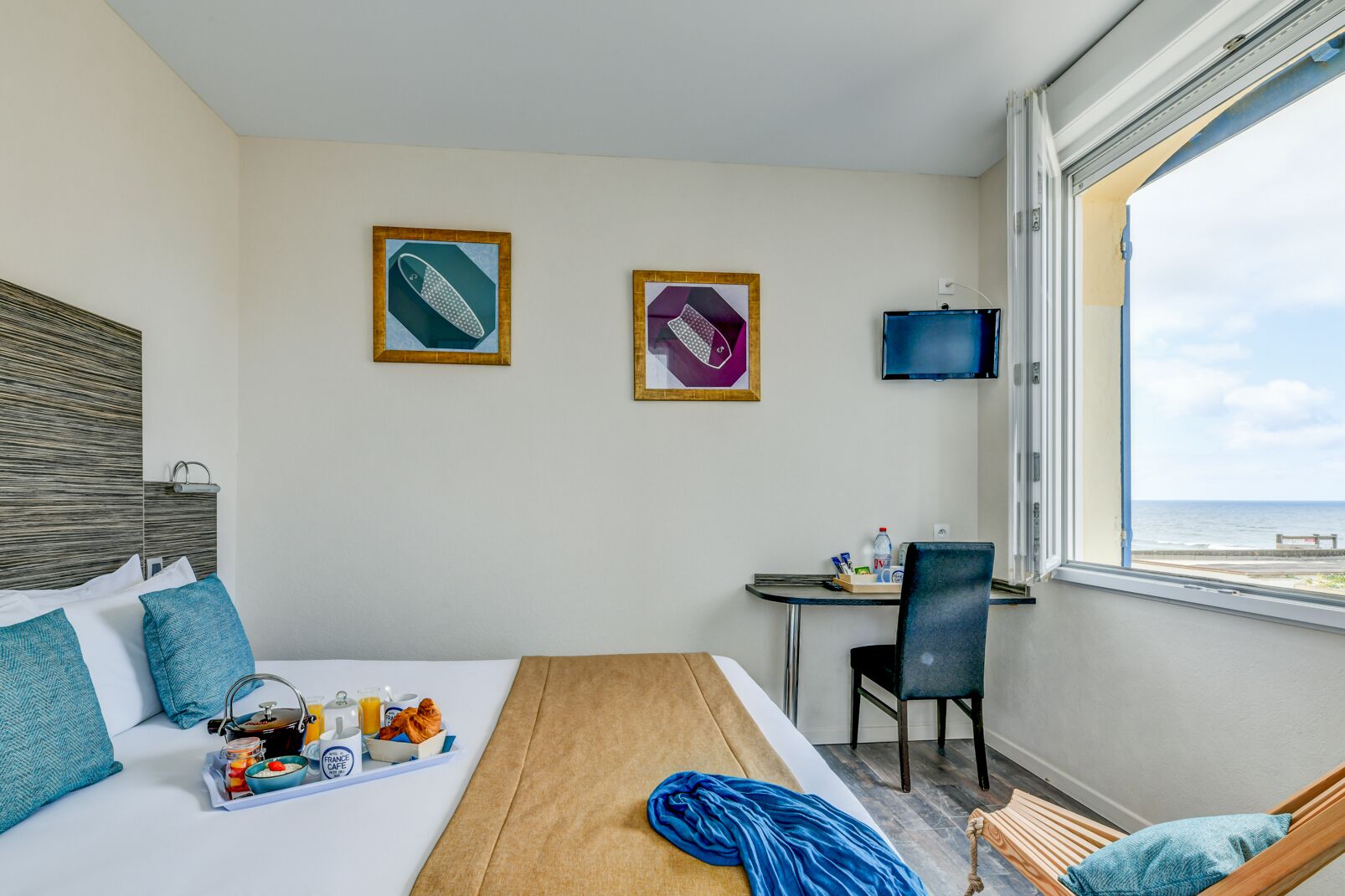A hotel room with a bed, a dining table with breakfast, a desk, a chair, a TV, and a view of the sea.