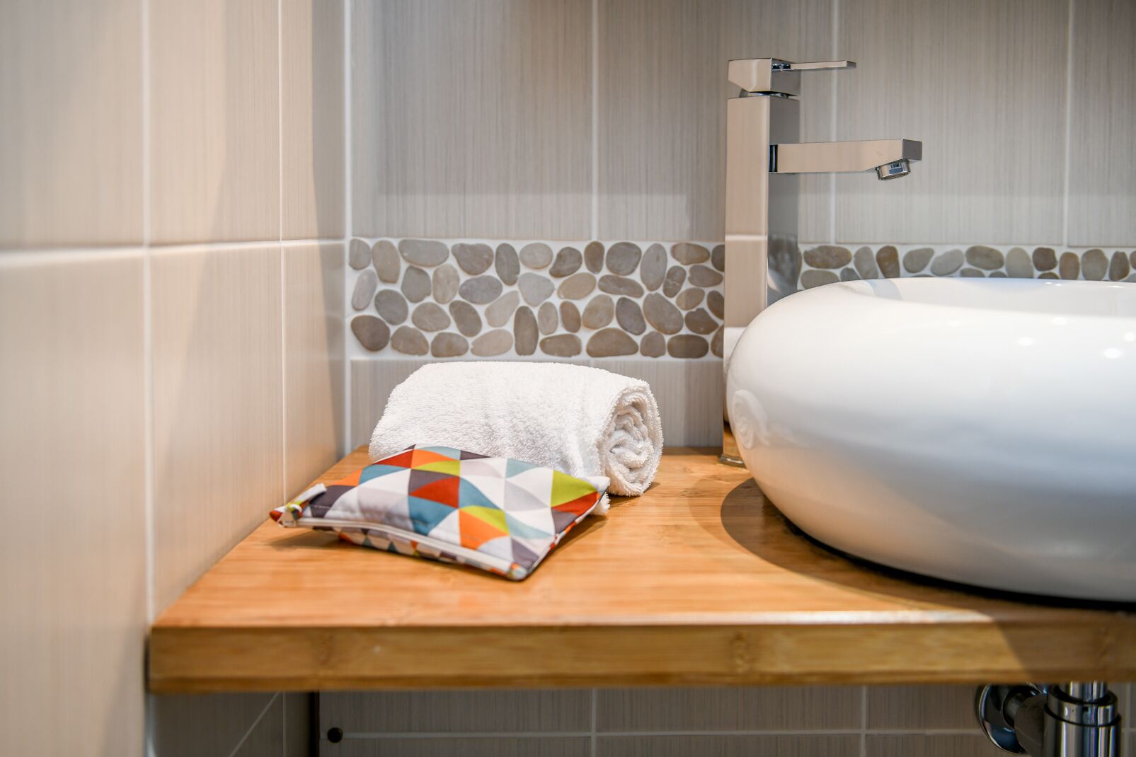 Un baño con una bañera blanca, un estante de madera con una toalla y un cojín de colores, y una pared con estampado de piedra.