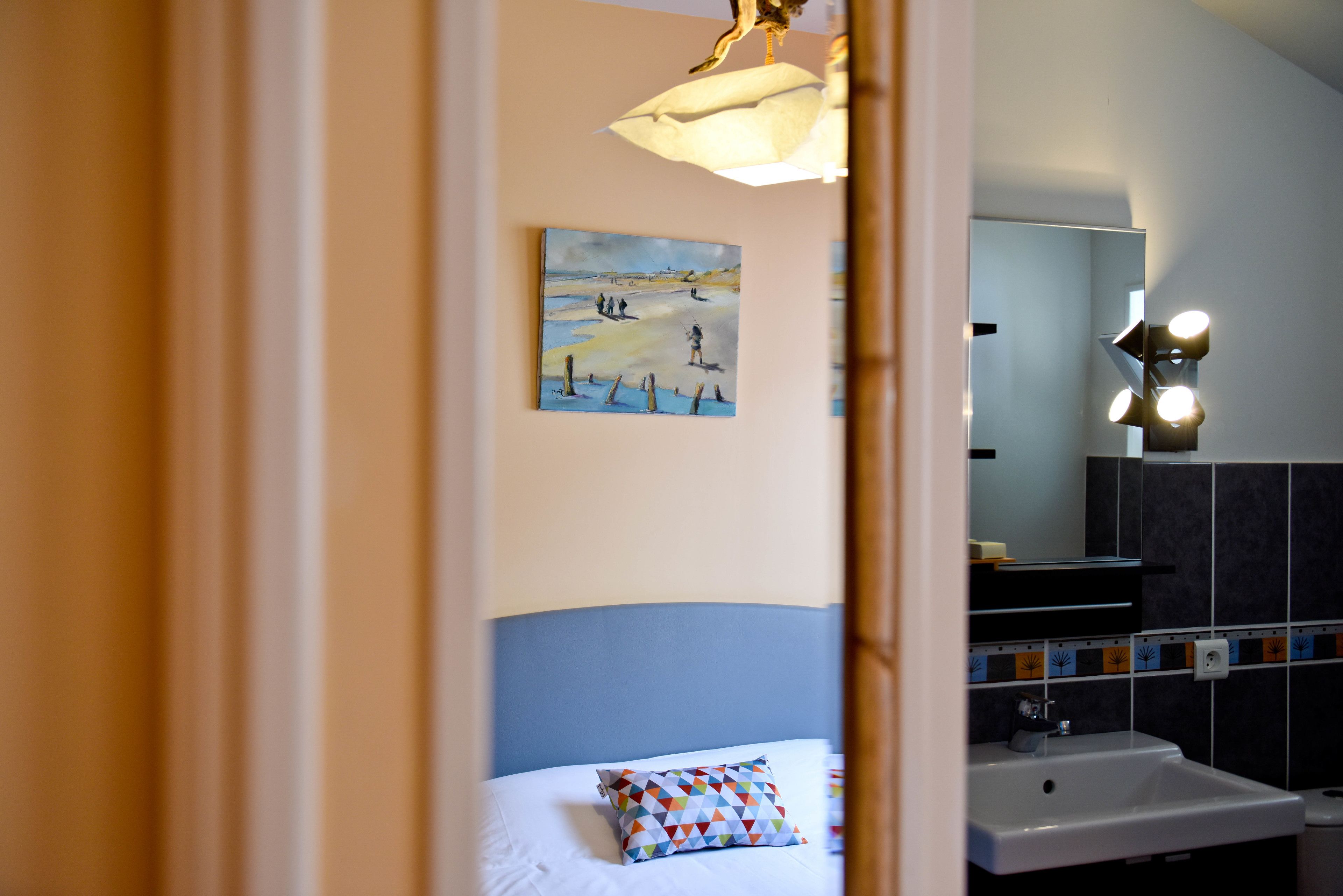 Une chambre d’hôtel avec un lit, un tableau au mur, une salle de bain avec un lavabo et un miroir, et un luminaire au-dessus du miroir.