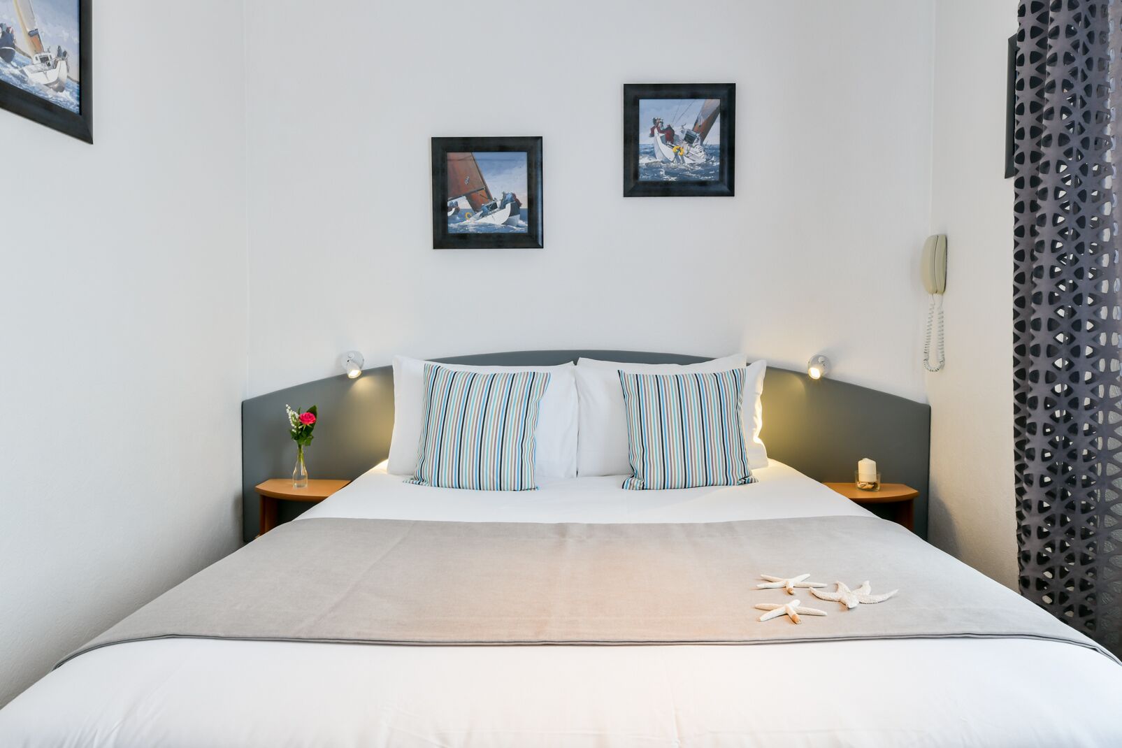 A hotel room with a bed, two bedside tables, a nightstand, a vase with flowers, and two framed pictures on the wall.