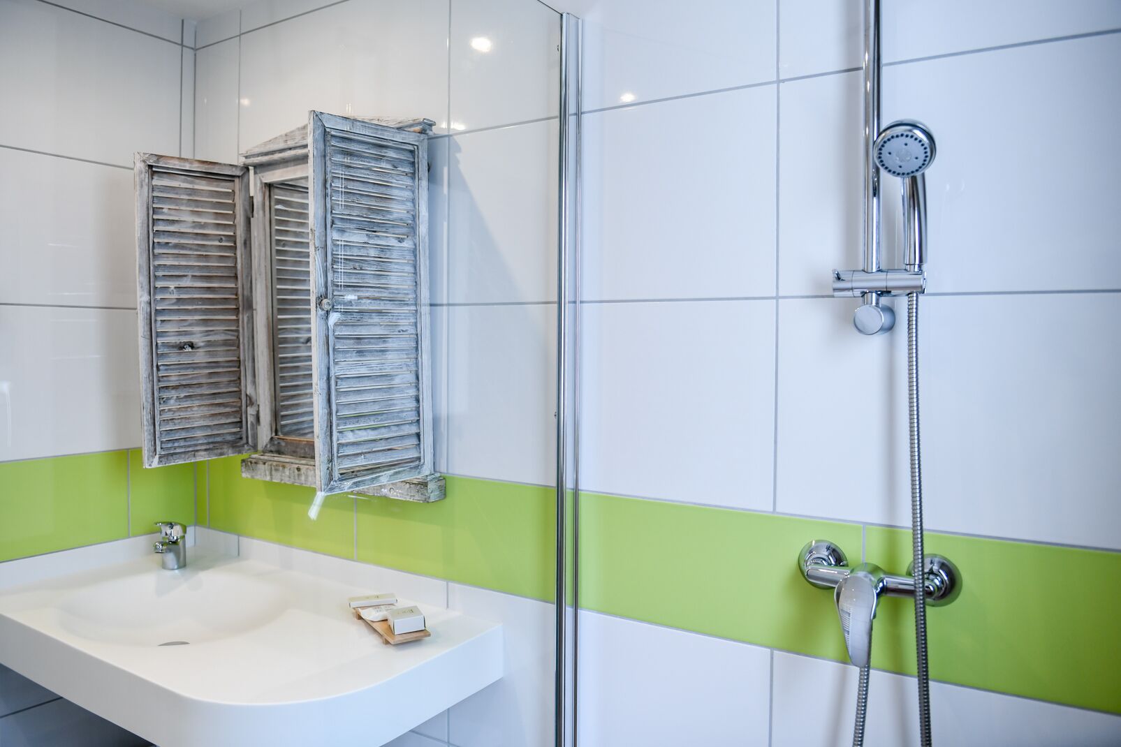 A bathroom with a white sink, green-tiled walls, a shower with a handheld showerhead, and a mirror with wooden shutters.