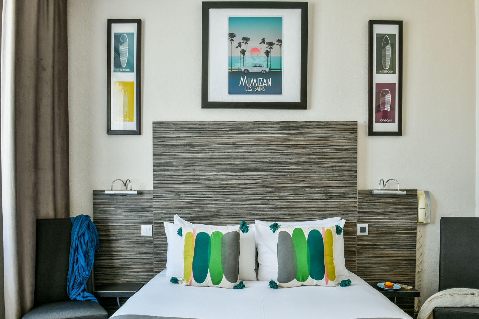 Quarto de hotel com uma cama grande, uma cabeceira de madeira, um cartaz emoldurado de Mimizan e uma pequena mesa com um prato e uma chávena.