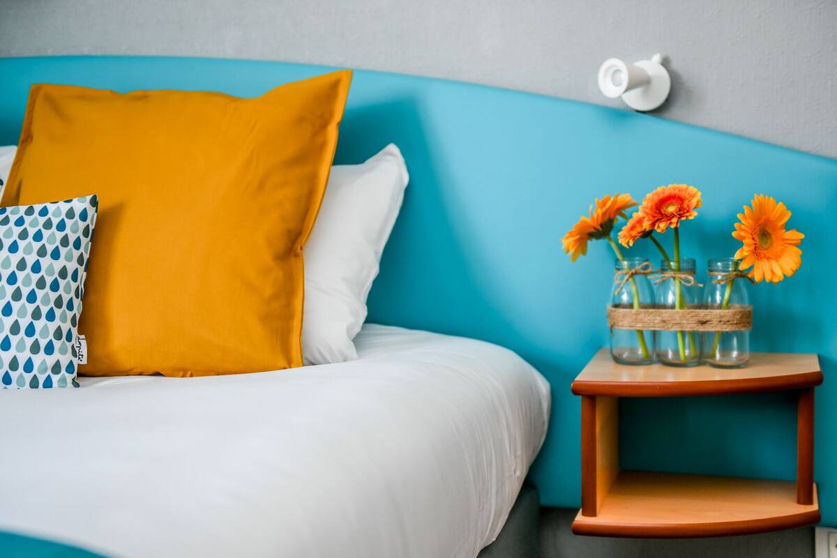 Una cama con ropa de cama blanca, un cojín amarillo mostaza y un cojín estampado en azul y blanco. Se observa un pequeño mesita de noche con un jarrón de flores anaranjadas.