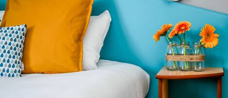 Un lit avec une literie blanche, un oreiller jaune moutarde et un oreiller à motifs bleus et blancs. On y trouve une petite table de chevet en bois sur laquelle est posé un vase rempli de fleurs oranges.