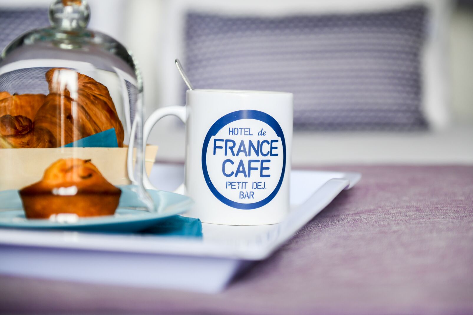 Eine weiße Tasse mit dem Schriftzug "HOTEL de FRANCE CAFE", ein Glas mit einem Muffin und ein Teller auf einem Tisch.
