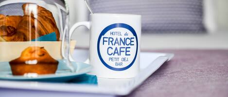 Una taza blanca con la leyenda "HOTEL de FRANCE CAFE", un vaso transparente con un muffin, y un plato azul sobre una mesa.