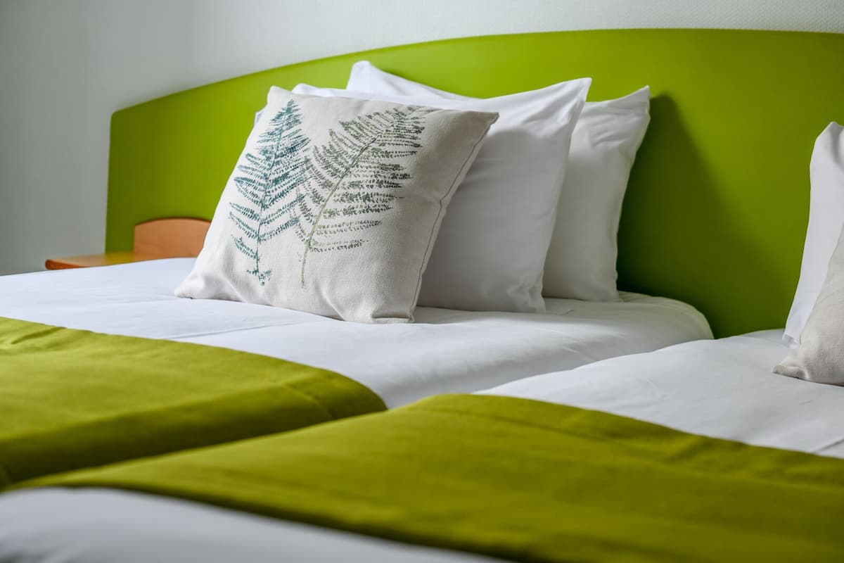 Una cama bien tendida con sábanas blancas y detalles verdes, un cabecero verde y una almohada con un diseño de helecho.
