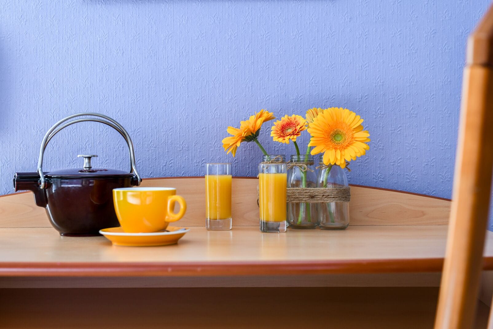 Una mesa de madera con una tetera, una taza amarilla, dos vasos de jugo de naranja y un florero con flores naranjas.
