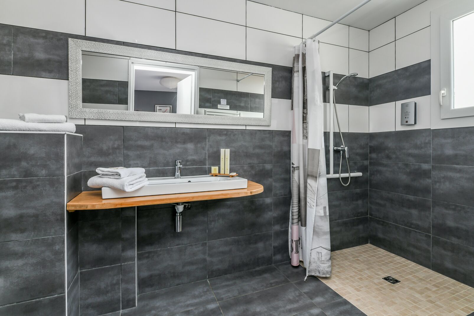 Une salle de bain moderne avec un grand miroir, un lavabo doté d’un plan de toilette en bois, une douche avec une porte en verre et des murs recouverts de carreaux foncés.