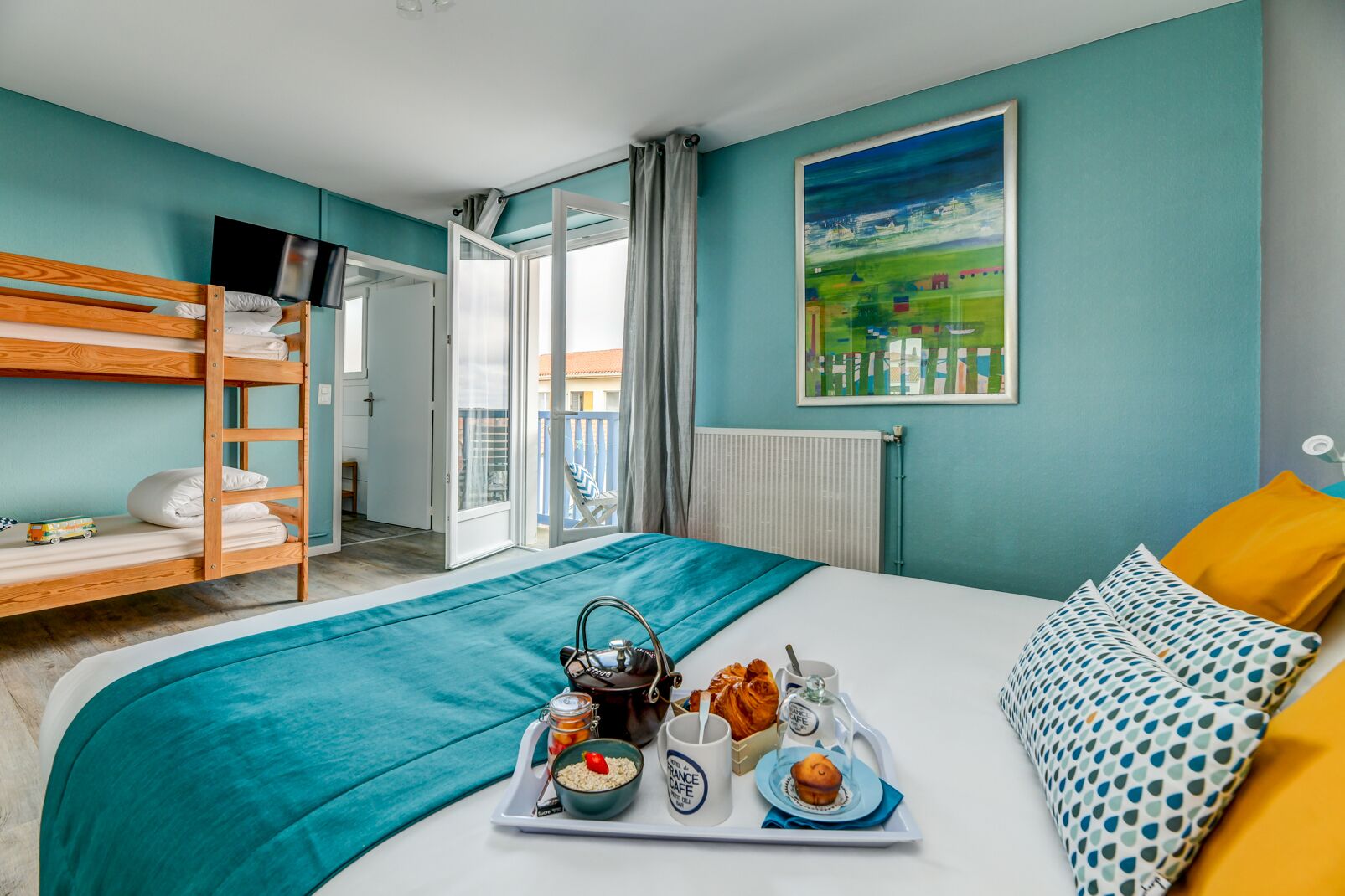 Habitación de hotel con literas, una cama grande con una bandeja de desayuno, un cuadro en la pared y vistas al balcón exterior.