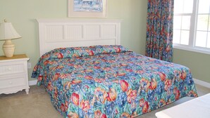 A bedroom with a floral bedspread, a white dresser, a lamp, a painting on the wall, and a window with curtains.