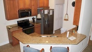 A kitchen with wooden cabinets, a stainless steel refrigerator, a microwave, and a sink. There are two bar stools and a hanging light fixture. - 3804 Bay Village (North Myrtle Beach)