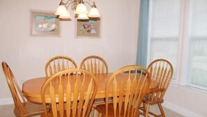 A dining area with a wooden table and chairs, framed artwork on the wall, and a window with blinds. - 3804 Bay Village (North Myrtle Beach)