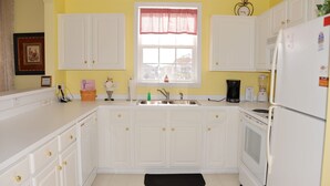 A kitchen with white cabinets, a black rug, a window with a red valance, and a clock on the wall. - 434 Ironwood (North Myrtle Beach)