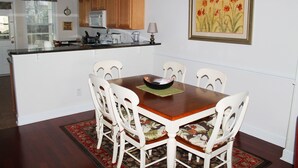 Un comedor con mesa de madera, sillas blancas, cocina al fondo y un cuadro con motivos florales en la pared.