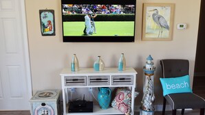 Una sala de estar con un televisor mostrando un partido de golf, una consola blanca decorada con objetos, un sillón con un cojín con motivos marinos y una mesita auxiliar con una radio.