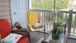 A porch with a white door, a yellow chair, a wicker chair, a small table with a plant, and a red cushion. - 522 Cypress Bend (North Myrtle Beach)