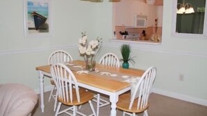A dining area with a wooden table and white chairs, a vase with flowers, and a kitchen in the background. - 522 Cypress Bend (North Myrtle Beach)