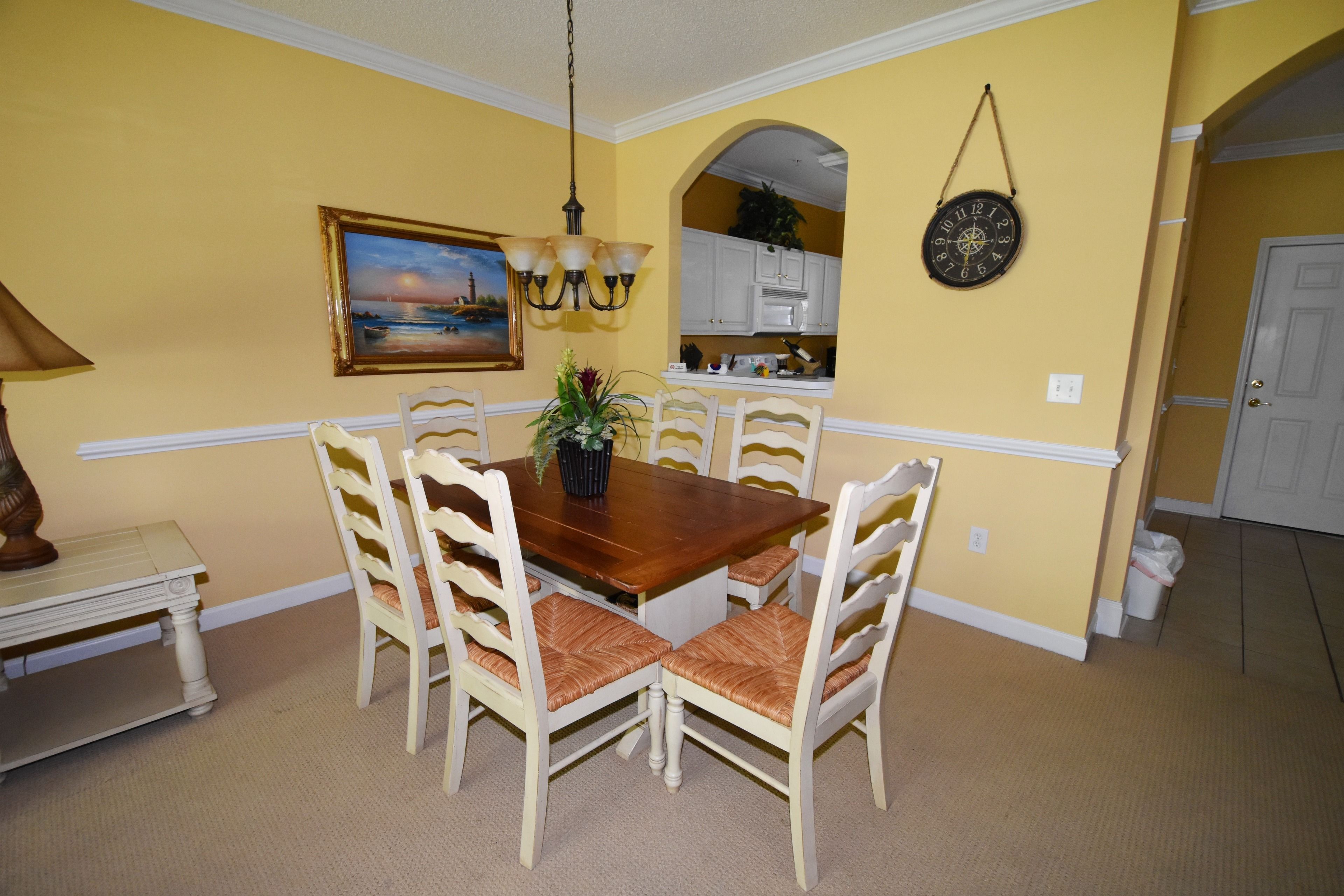 A dining area with a wooden table and white chairs, a painting on the wall, and a kitchen in the background.
