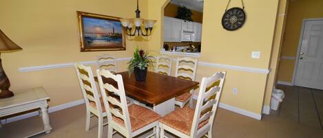 A dining area with a wooden table and white chairs, a painting on the wall, and a kitchen in the background.