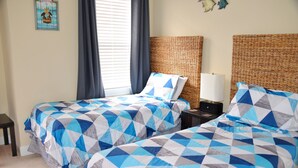 A room with two beds, each with a geometric-patterned bedspread, a wicker headboard, a bedside table with a lamp, and a framed welcome sign on the wall. - 327 The Havens (North Myrtle Beach)