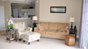 A living room with a beige sofa, patterned armchair, and ottoman. There are two side tables with lamps, a small side table with a plant, and a framed picture on the wall. - 327 The Havens (North Myrtle Beach)