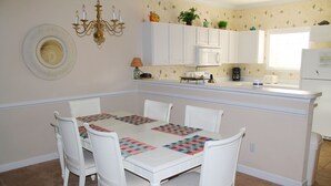 A dining area with a white table and chairs, a chandelier, and a kitchen in the background. - 622 Willow Bend (North Myrtle Beach)