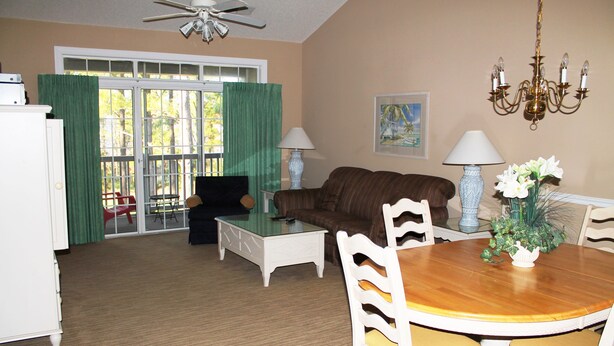 A living room with a brown sofa, a glass coffee table, a dining table with chairs, a chandelier, and a painting on the wall.