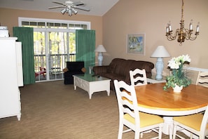 A living room with a brown sofa, a glass coffee table, a dining table with chairs, a chandelier, and a painting on the wall. - 633 Cypress Bend (North Myrtle Beach)