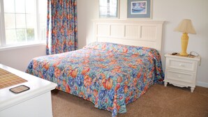 A bedroom with a floral bedspread, a white dresser, a nightstand with a lamp, and two framed pictures on the wall. - 713 Ironwood (North Myrtle Beach)