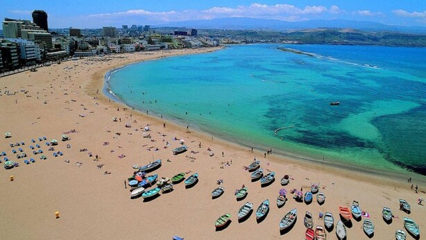 Una playa con numerosos barcos y sombrillas, un mar azul cristalino y un paisaje urbano al fondo.