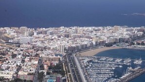 Vista aérea de una ciudad costera con una densa edificación urbana, un puerto deportivo con numerosos barcos y una carretera curva destacada que conduce a la playa.