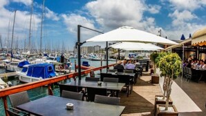 Un puerto deportivo con barcos amarrados, comedor al aire libre con sombrillas y vistas del horizonte urbano.