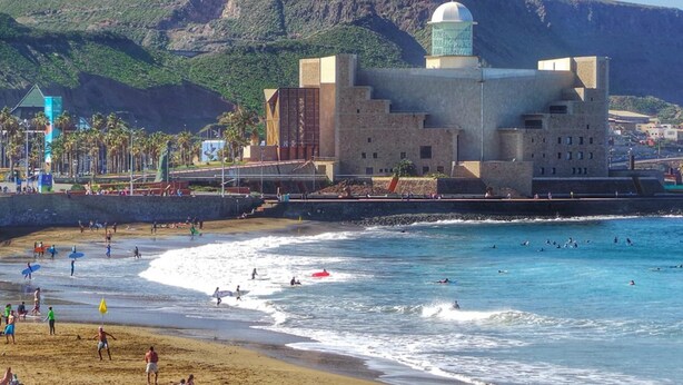 Un edificio costero con cúpula, junto a una playa donde hay gente surfeando y nadando.
