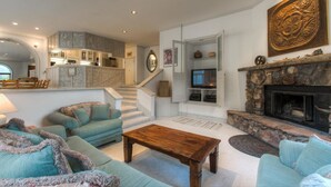 A spacious living room with a fireplace, a kitchen in the background, and a staircase leading to another level.