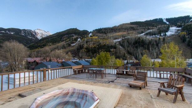 A wooden deck with outdoor seating and a hot tub, overlooking a mountainous landscape with snow patches and a river.