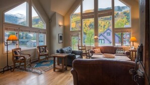 Ein geräumiges Wohnzimmer mit großen Fenstern, Holzböden und vielfältigen Sitzgelegenheiten, darunter eine Couch und Sessel.