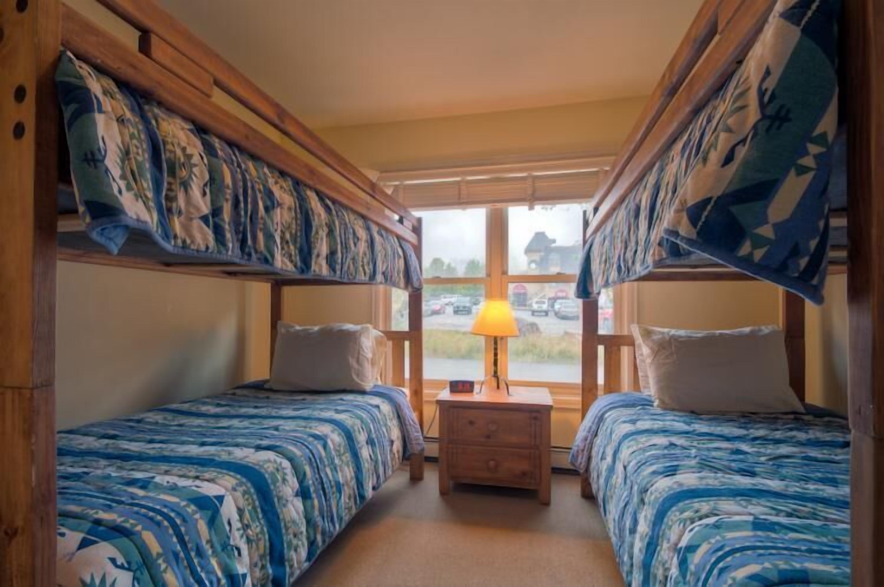 A room with two beds, each with a blue and white patterned bedspread, a small wooden nightstand with a lamp, and a window with a view of cars and a building outside.