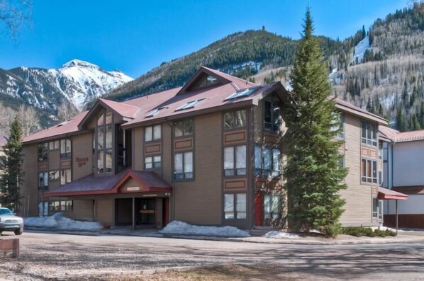 Un edificio de varios pisos con techo rojo, grandes ventanales y un paisaje montañoso al fondo.