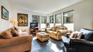 Ein geräumiges Wohnzimmer mit einem beigefarbenen Sofa, einem schwarzen Ledersofa und einem Couchtisch. Ein Fernseher steht auf einem Holzgestell, und ein großes Fenster bietet Blick ins Freie.
