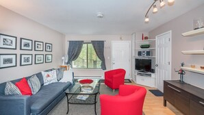 A modern living room with a grey sofa, red armchairs, a glass coffee table, and a white door leading to a kitchen.
