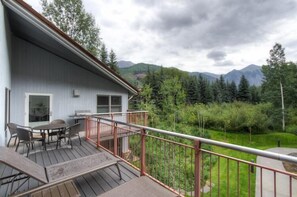 En træterrasse med et bord og stole, et lille udendørs køkken og udsigt til skov og bjerge.