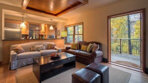 A modern living room with a sofa, a coffee table, and a fireplace. There is a large window with a view of trees. - Cascades A 3 3 Bedroom Condo By Accommodations in Telluride (Telluride)