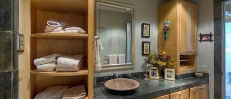 A bathroom with a wooden vanity, a sink with a bowl, and a mirror. There are towels on shelves and a vase with flowers on the counter.
