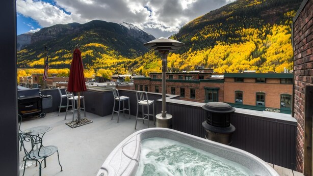 Een terras op het dak met een hottub, zitplaatsen buiten en uitzicht op glooiende bergen met herfstkleuren.
